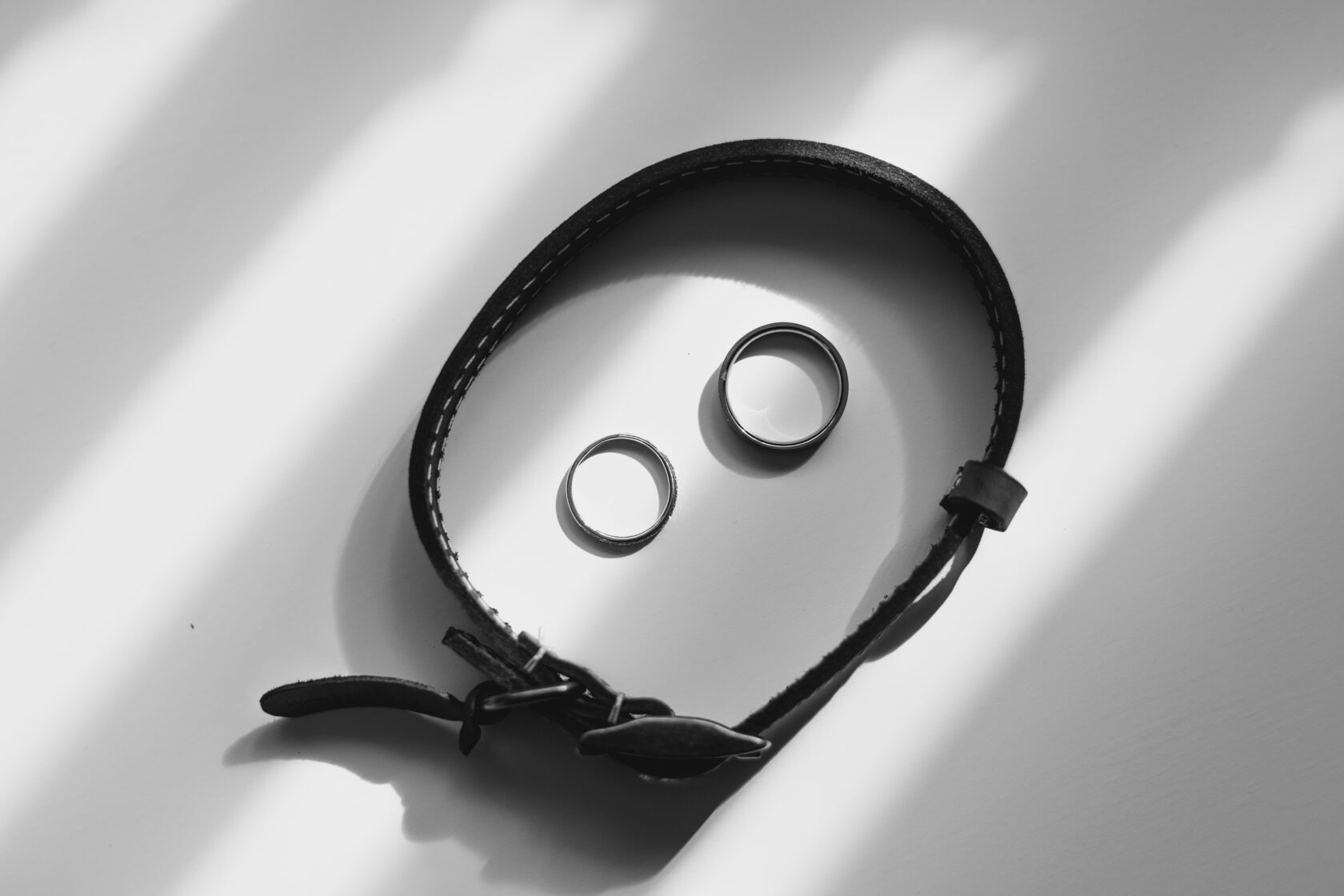 A close-up view of two metallic rings and a black leather bracelet placed on a light surface, with soft shadows creating a minimalist aesthetic. Shot for Lydia Turner.