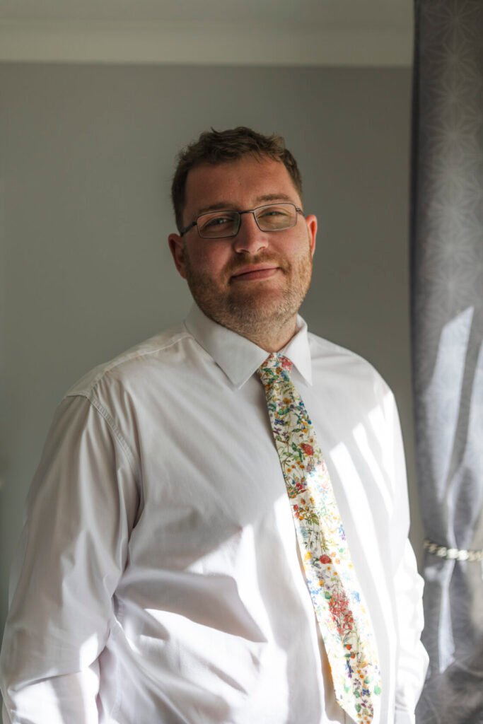 A man with glasses smiling, wearing a white shirt and a floral tie, standing in a well-lit room. Shot for Lydia Turner.