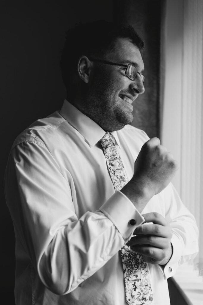 A man in a crisp white shirt and patterned tie smiles while fastening his cufflinks, looking out a window. Shot for Lydia Turner.