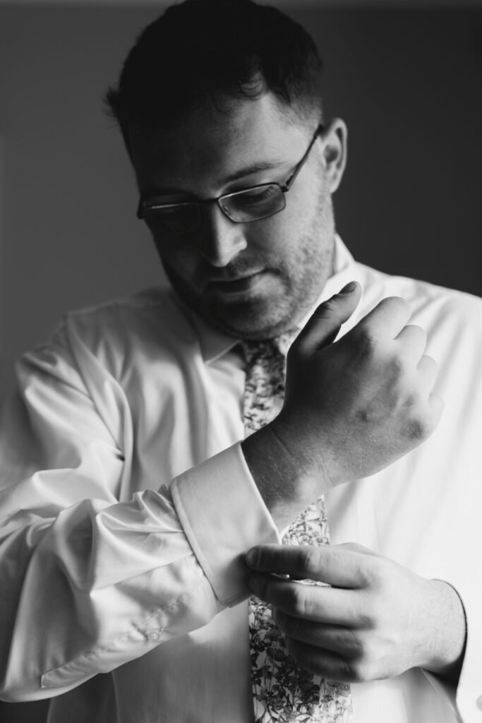 A man in a formal white shirt and patterned tie adjusts his cuff while looking down thoughtfully. Shot for Lydia Turner.