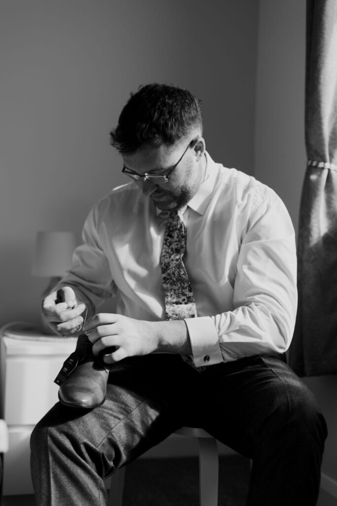 A man sits on a chair tying his shoelaces while wearing a dress shirt and patterned tie in a softly lit room. He appears focused and is preparing for a formal occasion. Shot for Lydia Turner.