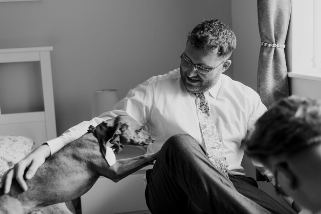 A man in a white shirt and patterned tie sits on a bed, laughing and interacting with a brown dog wearing a bow tie. The room has a warm atmosphere with light streaming through a window. Shot for Lydia Turner
