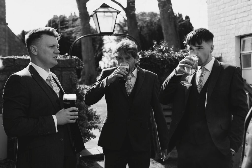 Three well-dressed men in suits standing outside, each holding a drink. One man in the centre is sipping a pint, while the others hold glasses. The scene is set in a sunny outdoor location with trees and a lantern in the background. Shot for Lydia Turner.