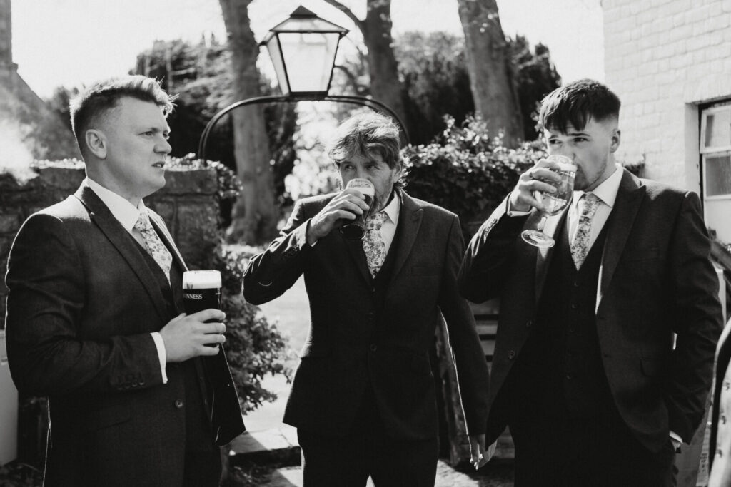 Three groomsmen in formal suits holding drinks outdoors, with one man drinking from a pint glass and another from a wine glass. They appear to be socialising in a sunny garden setting. Shot for Lydia Turner