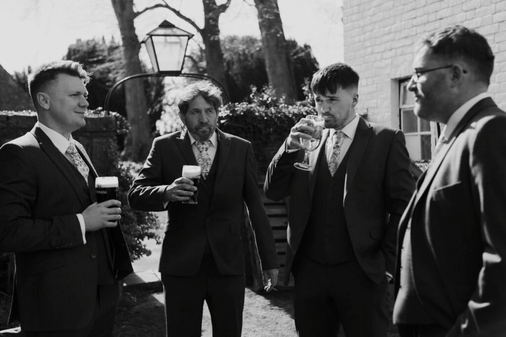 Four well-dressed men in suits holding drinks, standing outdoors in a sunny setting with a lamp post visible in the background. Shot for Lydia Turner