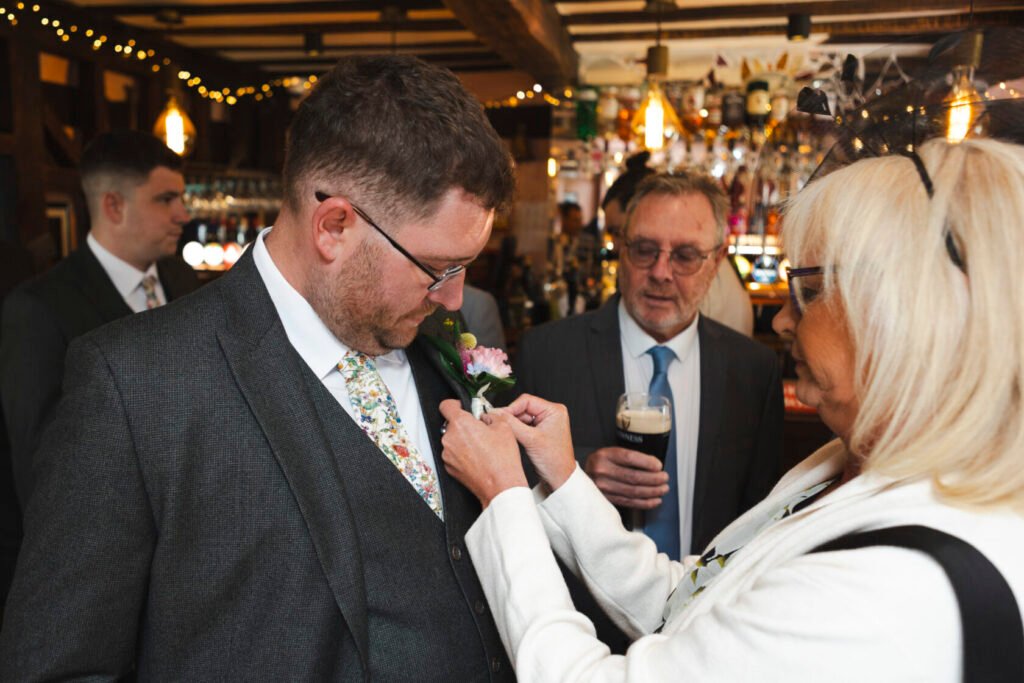 A man in a grey suit adjusts a floral boutonniere with the help of a woman in a white jacket. A man in a suit watches while holding a pint of Guinness. The background features a rustic pub interior with soft lighting and decorations.