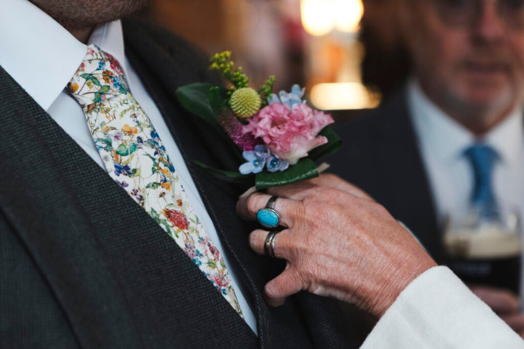 A close-up of a man wearing a floral tie and a boutonniere, while a hand adjusts the boutonniere. Shot for Lydia Turner