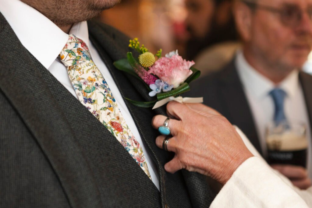 A close-up of a man's suit featuring a floral tie and a stylish boutonniere, while a hand gently adjusts the lapel. Shot for Lydia Turner.