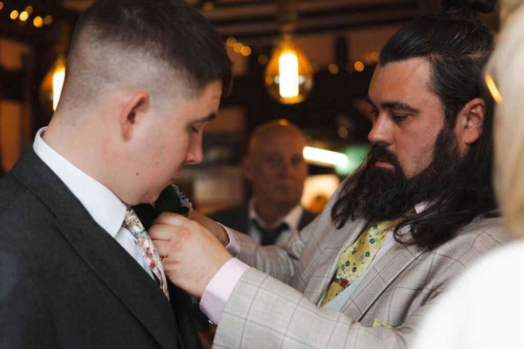 A groom in a dark suit receiving assistance from a groomsman in a plaid suit as he pins a flower onto his lapel. A third person can be seen in the background observing. Shot for Lydia Turner