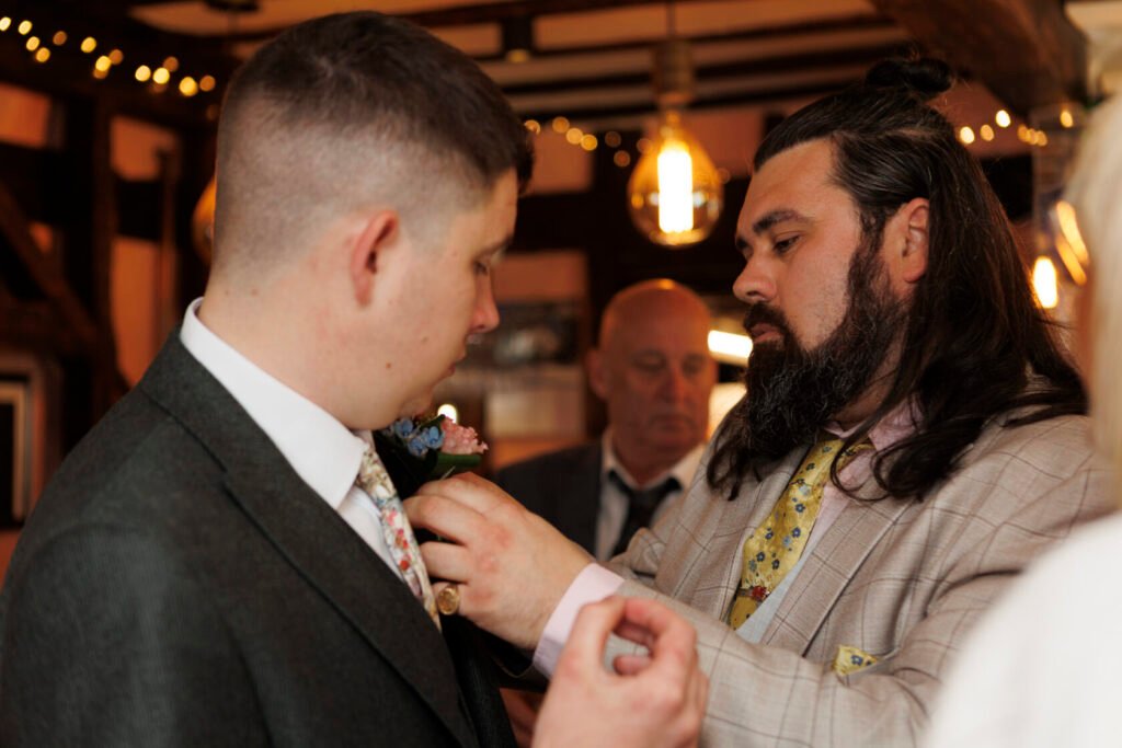 A groom in a grey suit having a floral boutonniere attached to his jacket by a friend in a light-coloured suit. Another man is visible in the background, all set in a rustic venue with warm lighting. Shot for Lydia Turner