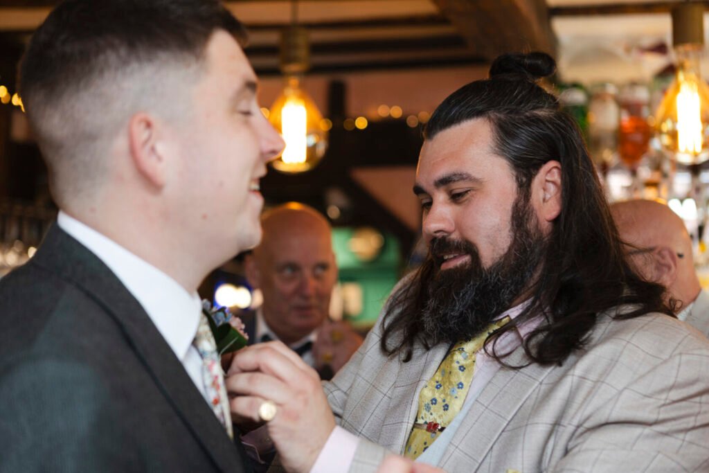 A joyful scene in a festive setting where a man with long hair and a beard is helping another man adjust a floral boutonniere on his suit. Shot for Lydia Turner.
