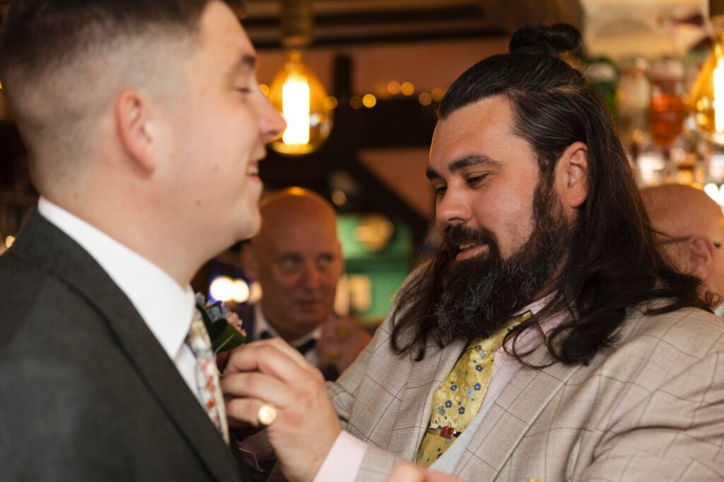 A man in a grey suit smiles as another man in a patterned tie adjusts a boutonniere on his lapel. A third man in the background watches with a smile. Shot for Lydia Turner.
