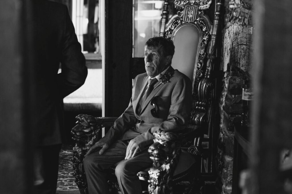 An elderly man sitting on an ornate throne with lion carvings, dressed in a grey suit and a blue patterned tie, lost in thought. A second person stands nearby, partially visible. Shot for Lydia Turner.