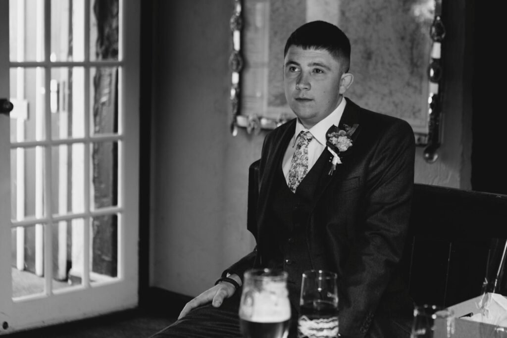 A young man in a formal suit sitting thoughtfully at a wedding reception. He is wearing a patterned tie and a floral boutonniere. Glasses of beer are on the table in front of him. Shot for Lydia Turner.