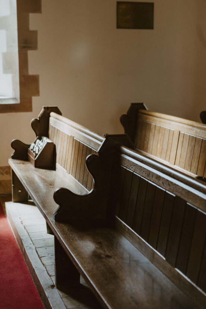 Close-up of wooden church benches with ornate ends, bathed in natural light from a nearby window. A box rests on one of the benches, likely for collecting items. Shot for Lydia Turner.