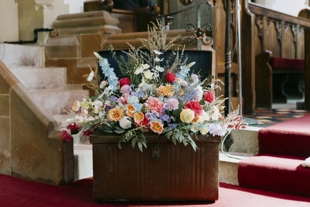 Vibrant floral arrangement featuring roses, daisies, and seasonal blooms in a metal container, placed on steps in a church setting. Shot for Lydia Turner
