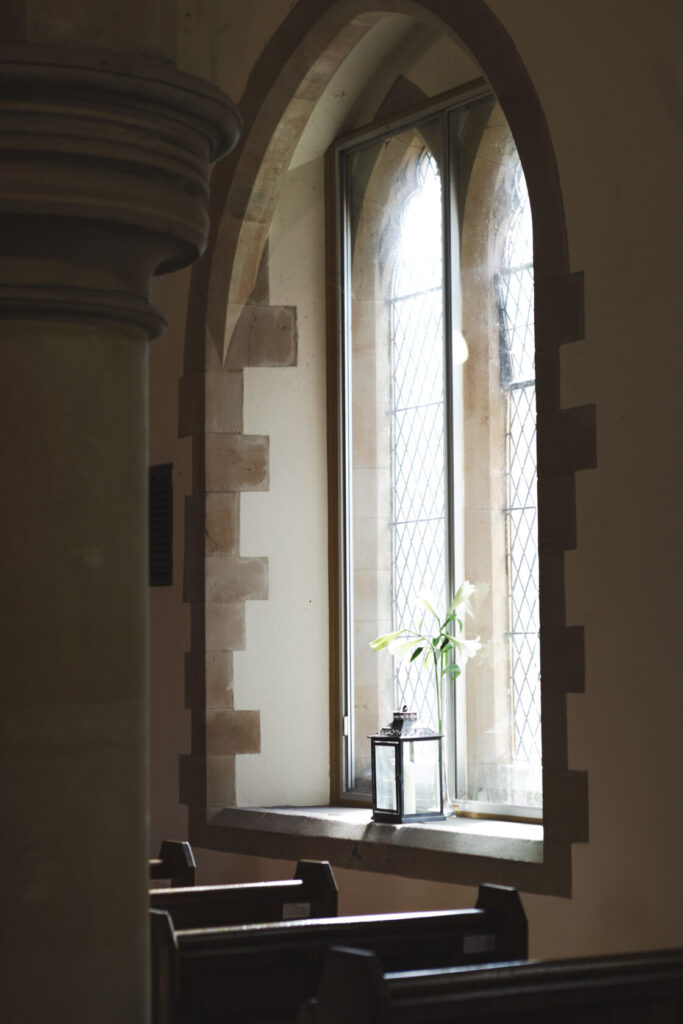 A sunlit church interior featuring a tall window with a small plant and a lantern on the sill, alongside a stone column and wooden pews. Shot for Lydia Turner.
