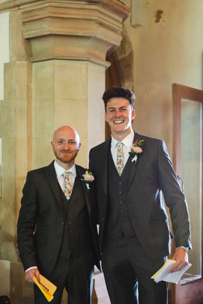 Two men in formal attire, one with a bald head and ginger beard, the other with dark hair, both wearing floral ties and boutonnières. They are smiling and standing in a hall with stone pillars. Shot for Lydia Turner