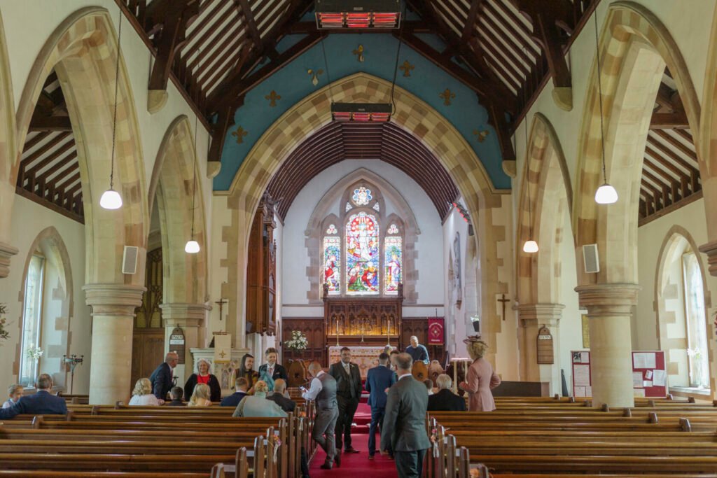 Wide view of a church interior featuring wooden pews, arched ceilings, and vibrant stained glass windows. Guests are gathered, engaging in conversation. Shot for Lydia Turner.