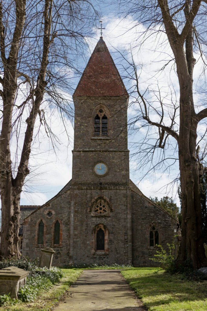 A stone church with a pointed steeple, clock on the side, surrounded by bare trees and gravestones, set on a grassy path. Shot for Lydia Turner