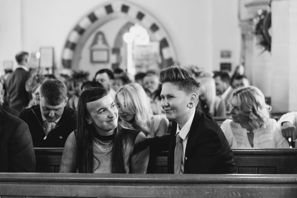 Two women share a joyful moment while sitting together in a church pew, surrounded by guests. The woman on the right, dressed in a suit, gently rests her hand on the shoulder of the woman on the left, who is wearing a black top. The scene is lively with guests in the background, all attending a wedding ceremony. Shot for Lydia Turner.