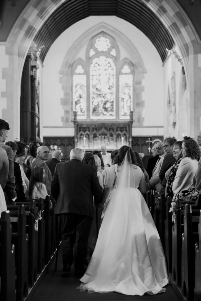 A bride in a white dress walks down the aisle of a church, holding hands with a man, amidst an audience of guests in formal attire. Stained glass windows and wooden pews are visible in the background. Shot for Lydia Turner.