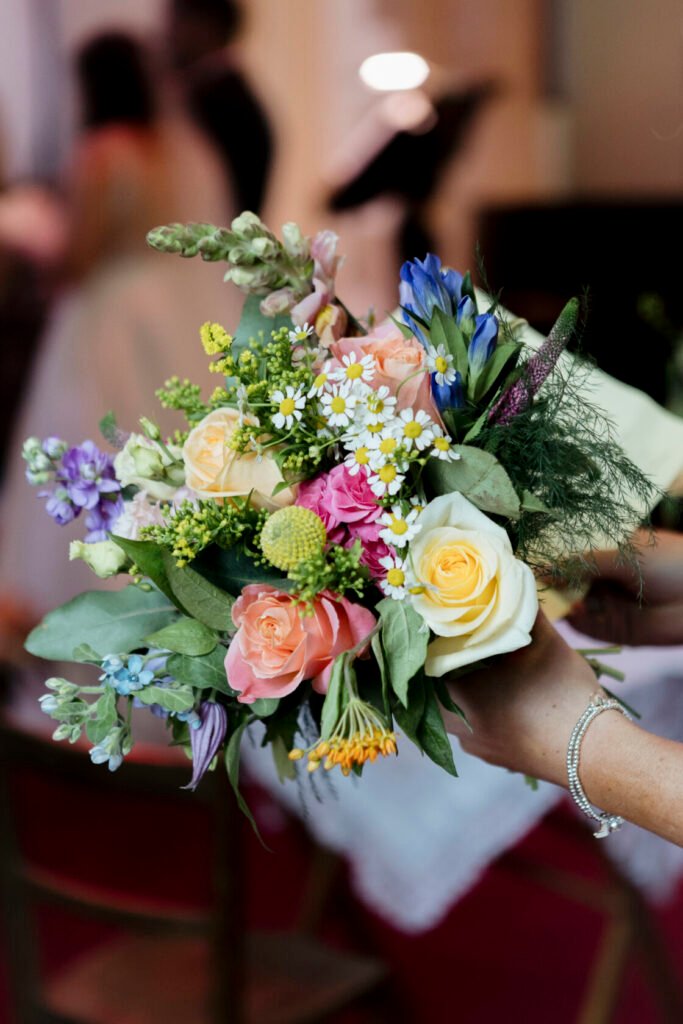 A vibrant wedding bouquet featuring various flowers including roses in shades of pink and yellow, daisies, and blue blooms. The bouquet is held by a person wearing a bracelet. Shot for Lydia Turner.