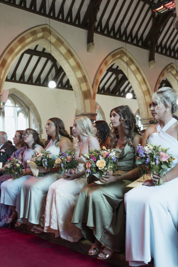 A group of six bridesmaids in elegant dresses, holding colourful bouquets, seated during a wedding ceremony inside a church with wooden arches and soft lighting. Shot for Lydia Turner.