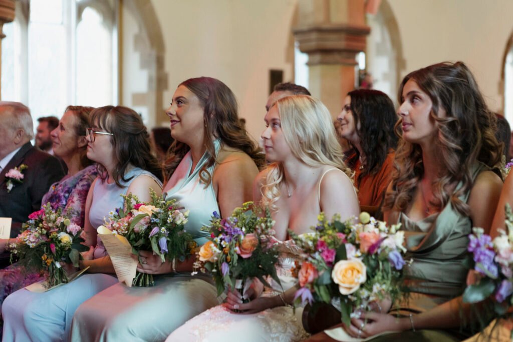 A group of bridesmaids sitting during a wedding ceremony, holding bouquets of flowers and wearing elegant dresses in various pastel colours. Shot for Lydia Turner.