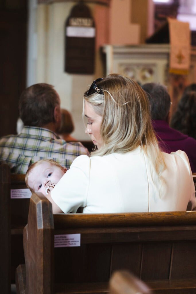 A woman with long, blonde hair, wearing a light-coloured dress, cradles a baby in her arms, who is looking curiously towards the camera. The background features people sitting in church pews.