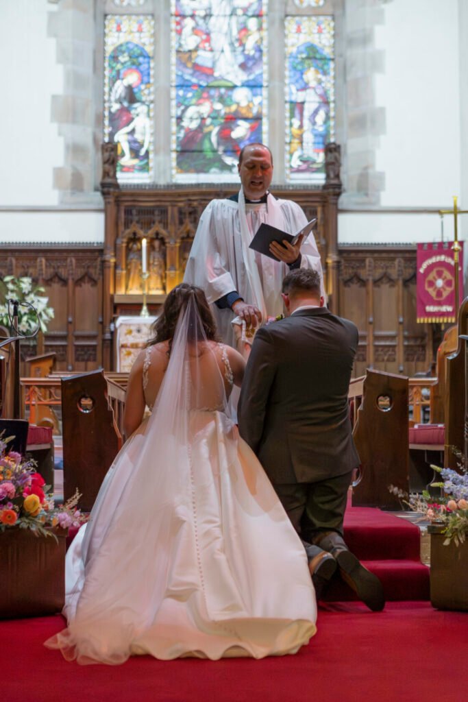 A bride and groom kneel before an officiant in a beautiful church, surrounded by stained glass windows and floral arrangements. The bride has a flowing veil and ornate gown, while the groom wears a dark suit. Shot for Lydia Turner.