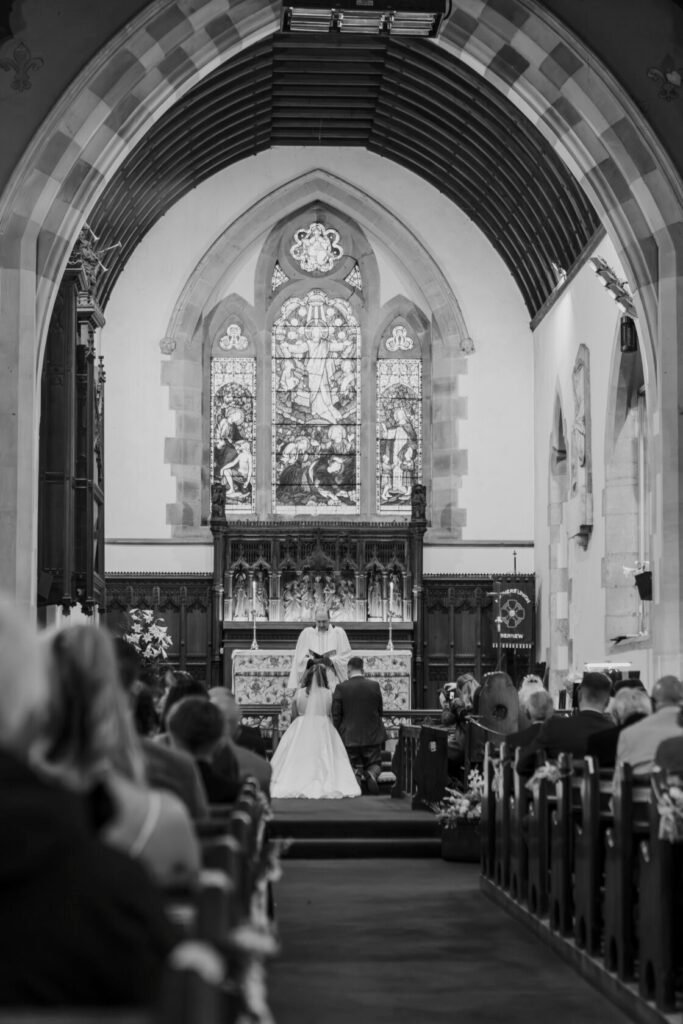 A bride and groom kneel at the altar during their wedding ceremony in a historic church, surrounded by stained glass windows and a gathering of guests. Shot for Lydia Turner