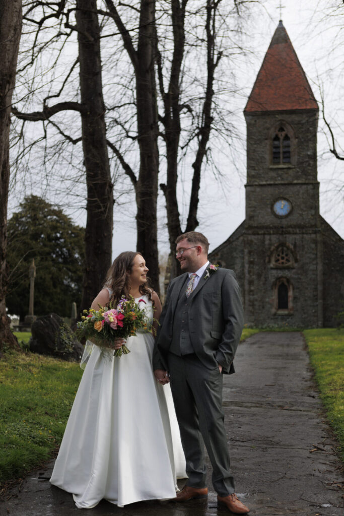 A smiling couple stands together outside a stone church, the bride wearing a flowing white gown and holding a vibrant floral bouquet, while the groom is dressed in a grey suit. The scene is framed by bare trees and a clock tower in the background. Shot for Lydia Turner.