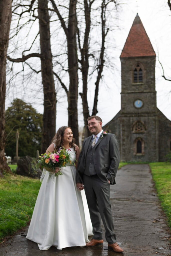 A smiling bride in a white dress holds a vibrant bouquet, standing beside a groom in a grey suit outside a historic church with a clock tower, surrounded by bare trees.