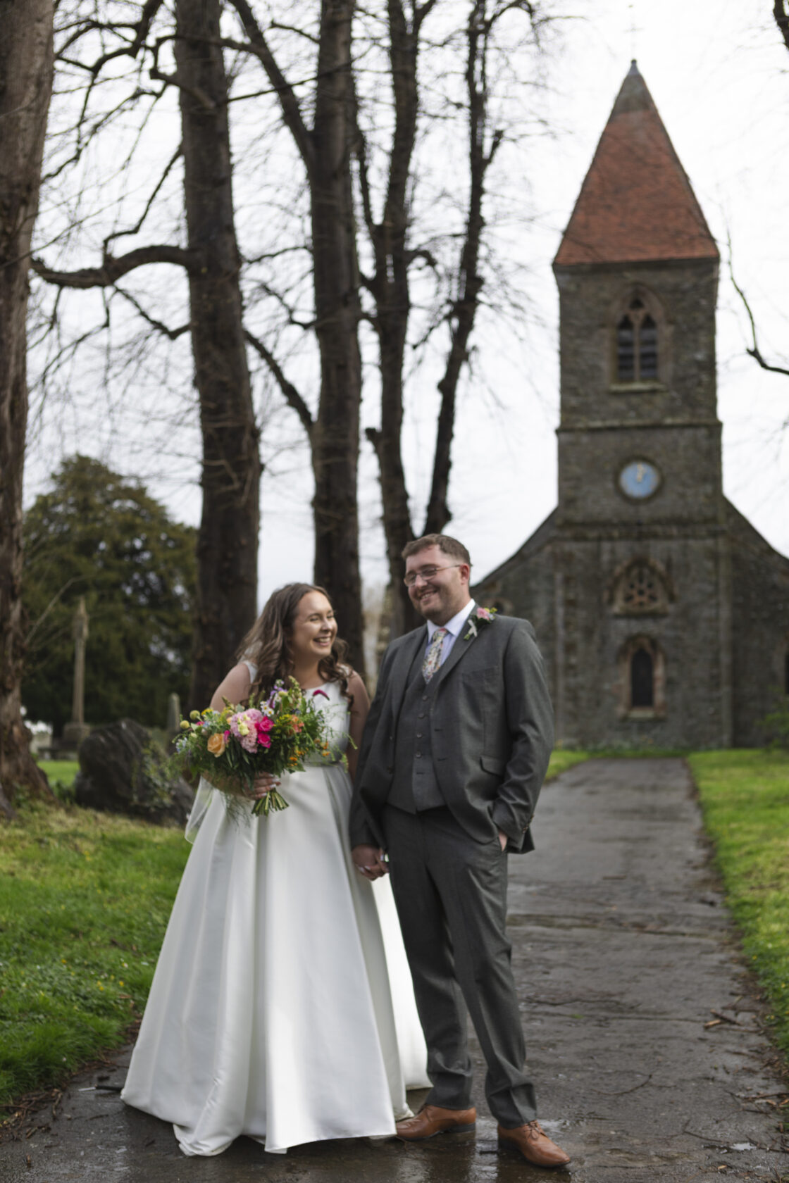 A smiling bride in a white dress holds a vibrant bouquet, standing beside a groom in a grey suit outside a historic church with a clock tower, surrounded by bare trees.