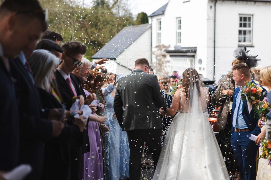 A couple exits a venue surrounded by guests throwing colourful confetti. The bride, wearing a white dress with a veil, and the groom in a dark suit walk amidst smiling friends and family. Shot for Lydia Turner.