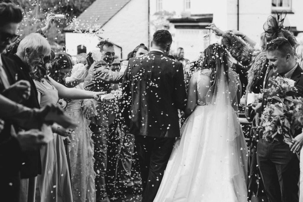 A bride and groom walk through a crowd of guests tossing confetti, surrounded by joyous family and friends during a wedding celebration. The scene is captured in black and white.