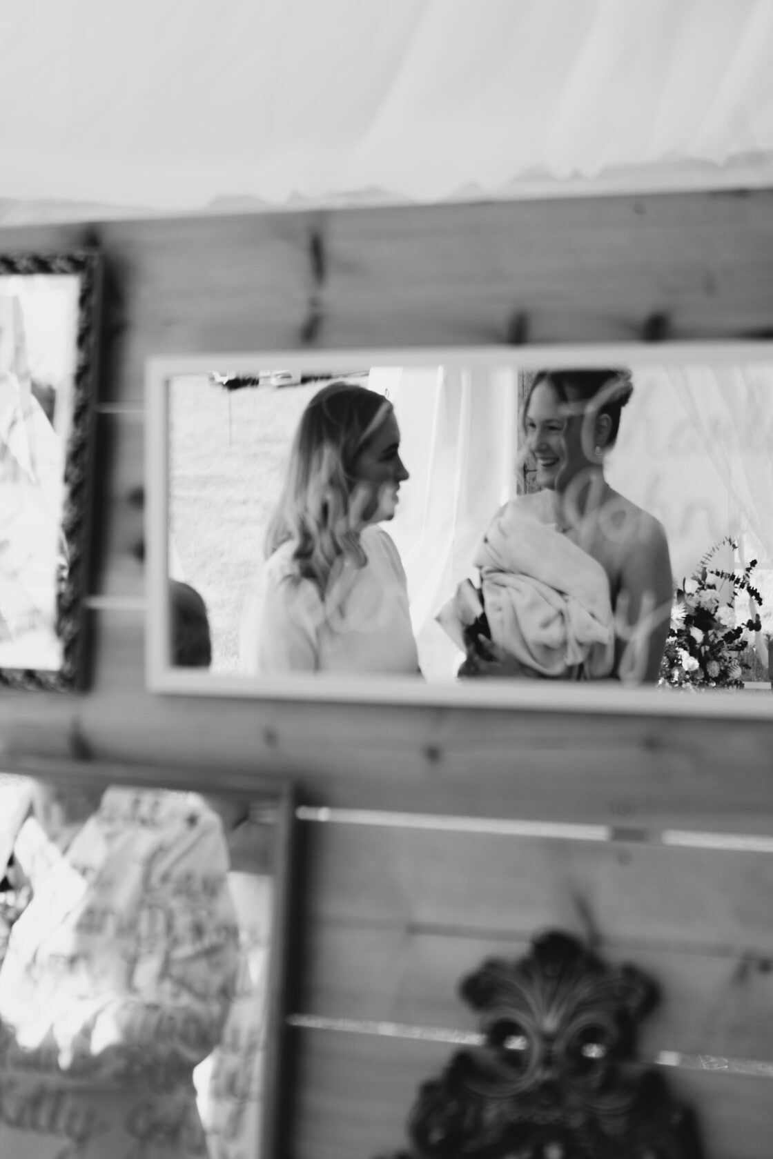 Two women, one in a strapless dress and the other in a white blouse, share a joyful moment reflected in a mirror. Flowers and decorations are visible in the background. Shot for Lydia Turner.