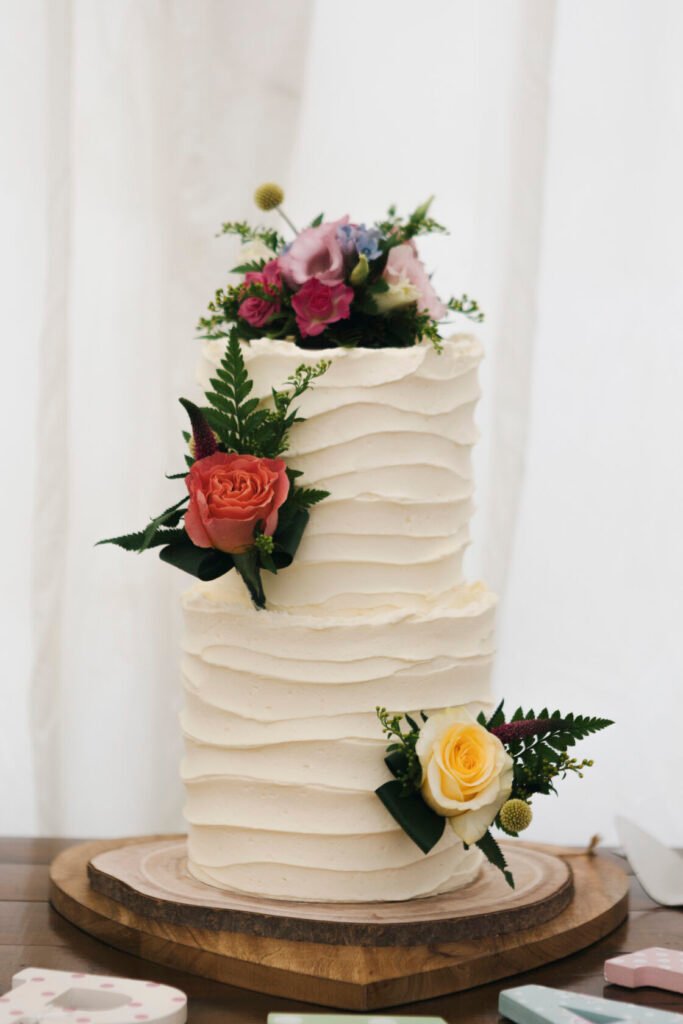 A beautiful two-tier wedding cake featuring textured white frosting and adorned with fresh flowers including pink and yellow roses, and greenery. Shot for Lydia Turner.
