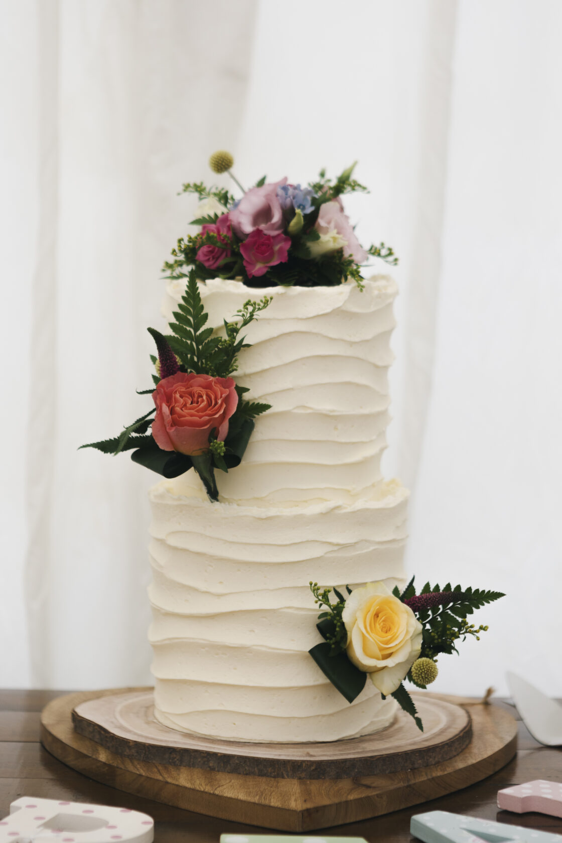A beautiful two-tier wedding cake featuring textured white frosting and adorned with fresh flowers including pink and yellow roses, and greenery. Shot for Lydia Turner.