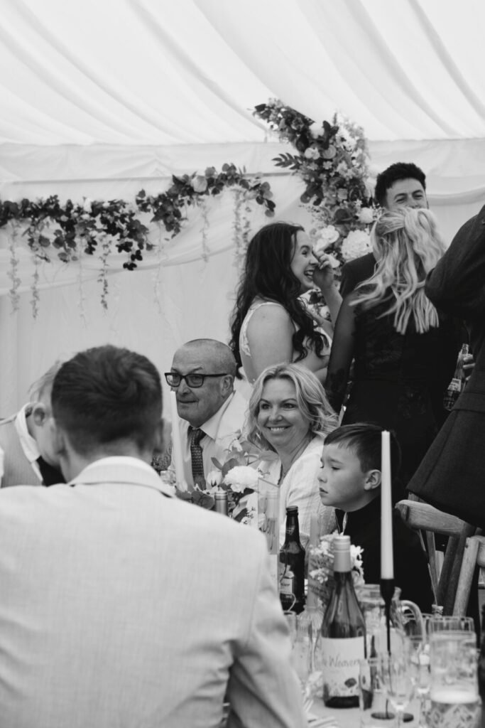A lively wedding reception with guests smiling and chatting, featuring a man in glasses, a cheerful woman, and a young boy sitting at the table. Floral decorations adorn the marquee. Shot for Lydia Turner.