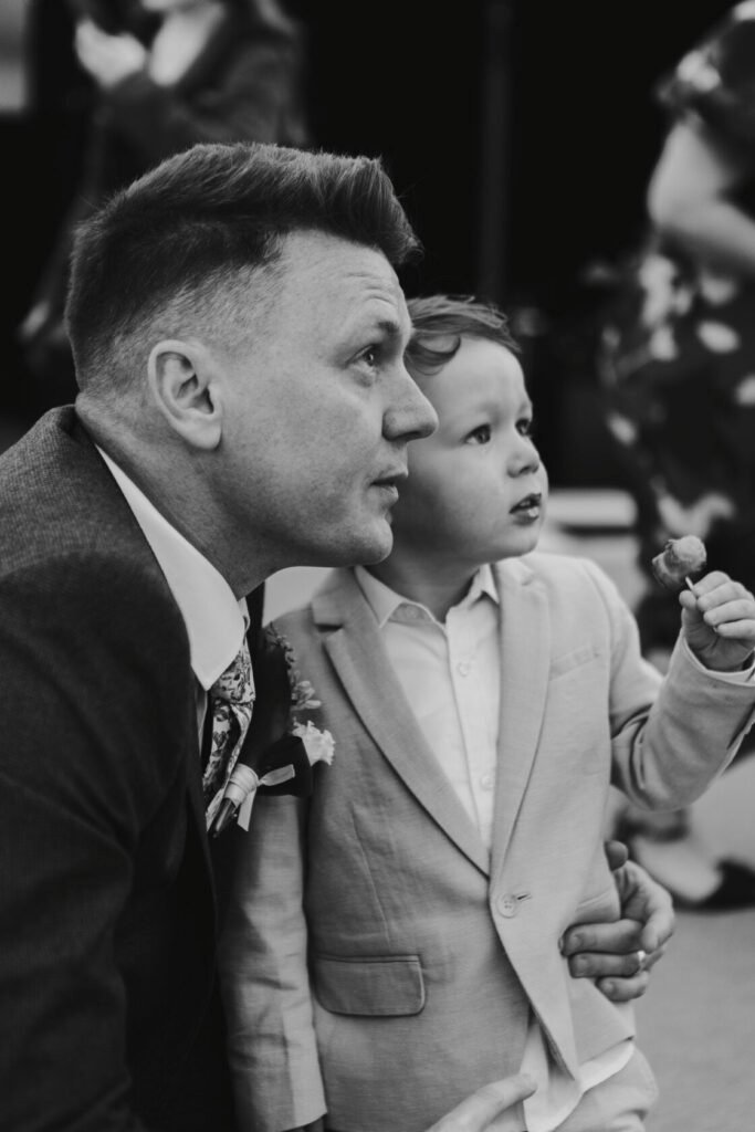 A man and a young boy sit close together, both appearing thoughtful. The man wears a dark suit with a floral tie, while the boy is dressed in a light-coloured suit, holding a piece of food. Shot for Lydia Turner.