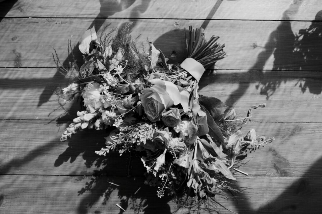 A black and white photograph of a flower bouquet featuring roses, greenery, and delicate blossoms, arranged with care, resting on a wooden surface. Shot for Lydia Turner.