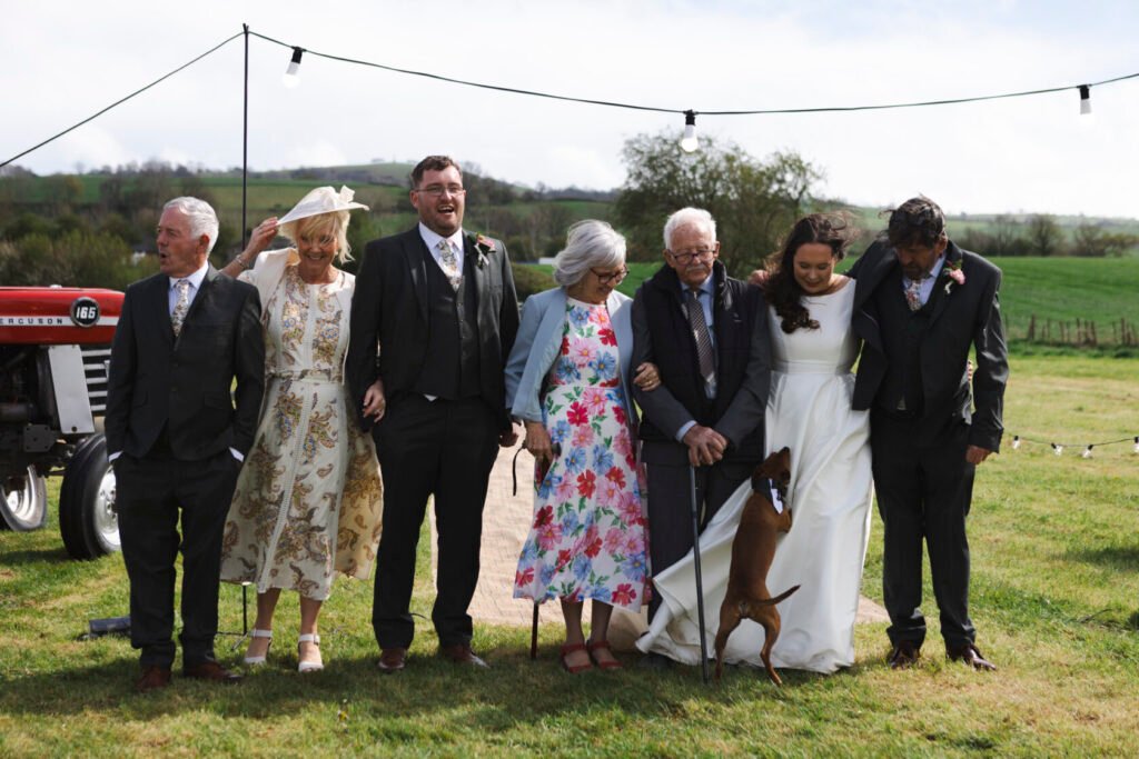 A joyful group of family members dressed in wedding attire, standing outdoors with a vintage tractor in the background. They include elderly individuals, adults, and a dog, all smiling and enjoying the moment. Shot for Lydia Turner.