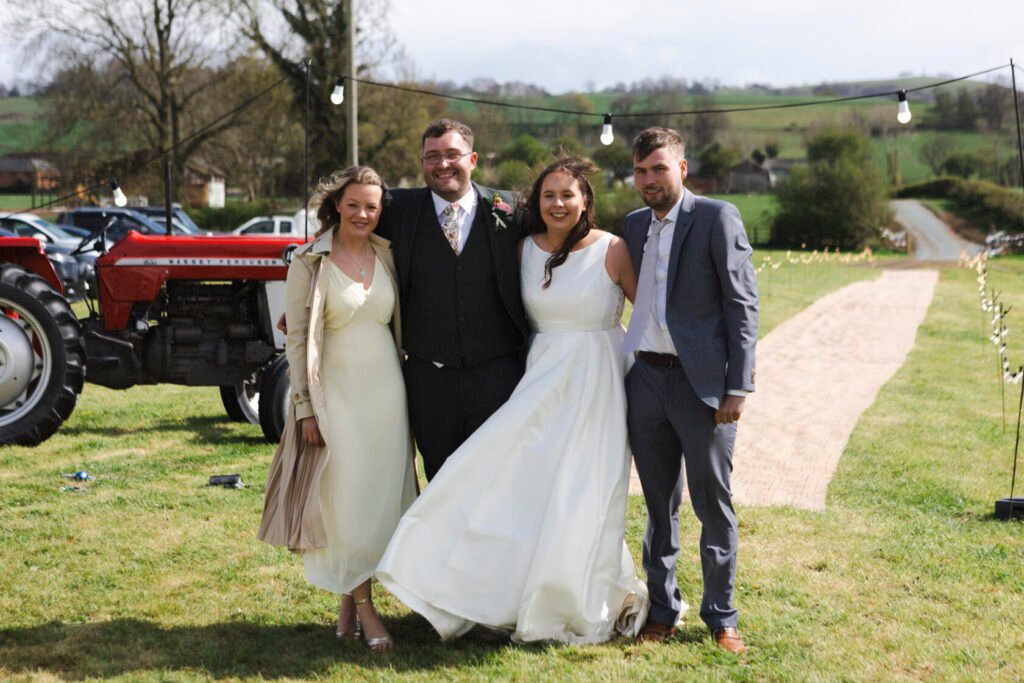A joyful gathering of four friends at a wedding, with one woman in an elegant white wedding dress, another in a light-coloured dress, and two men in formal attire, set against a scenic outdoor background with a red tractor and a rustic pathway.