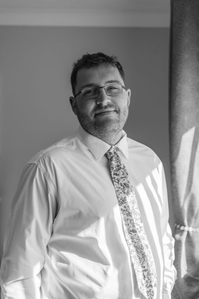 A man with glasses stands smiling confidently, dressed in a white shirt and patterned tie, with sunlight casting shadows behind him. Shot for Lydia Turner