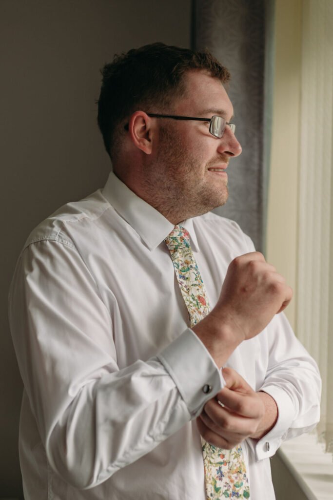 A man dressed in a white shirt with a colourful floral tie adjusts his cuff while looking out a window, showcasing a moment of anticipation. Shot for Lydia Turner.