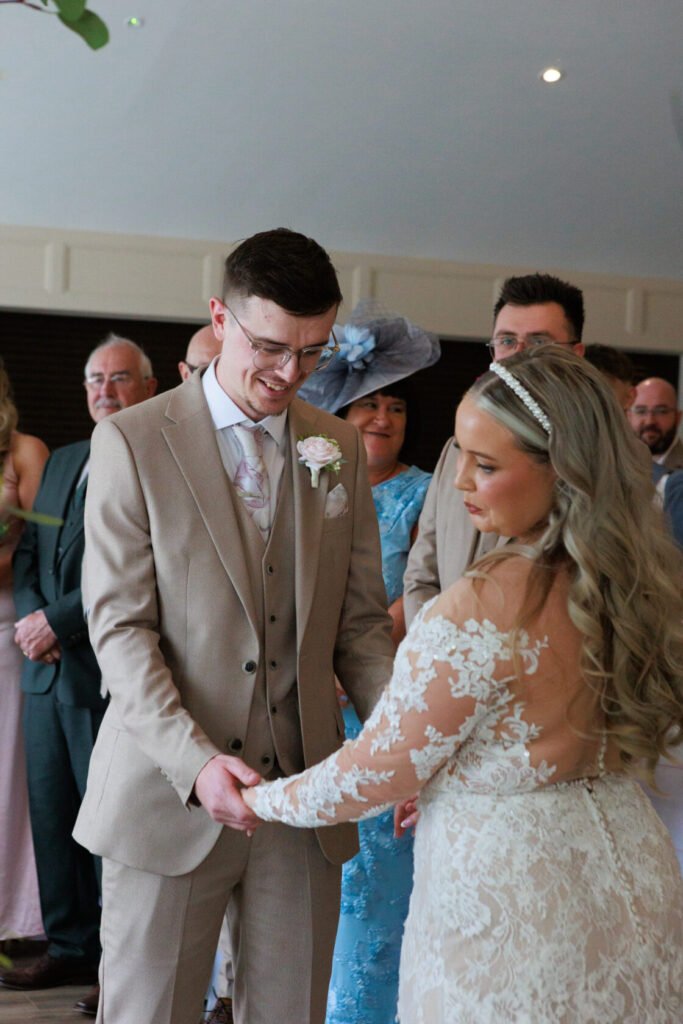 A groom in a light beige suit and bride in a lace wedding dress hold hands during a wedding ceremony, surrounded by smiling guests.