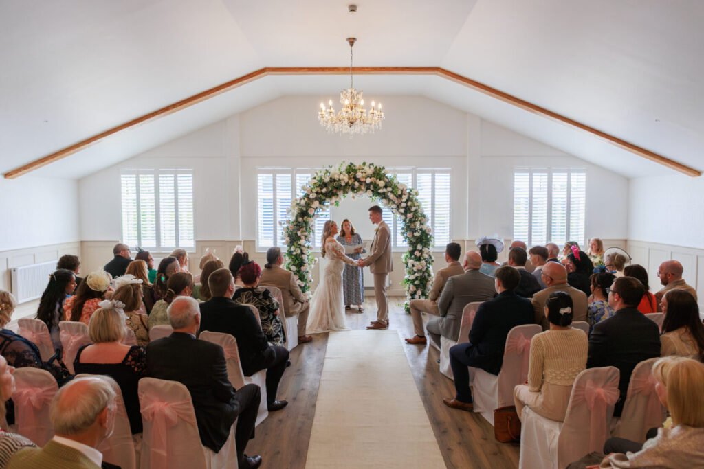A couple standing at the altar during their wedding ceremony beneath a floral arch, surrounded by seated guests in formal attire.