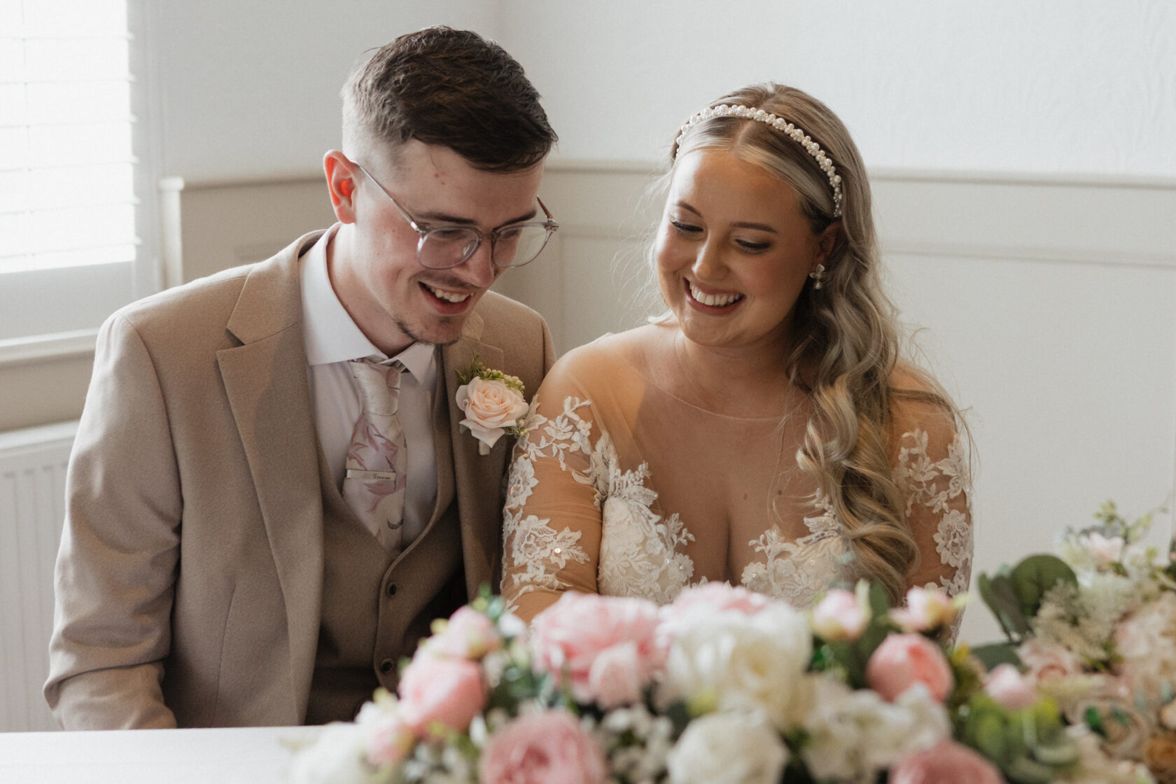 A smiling bride and groom sitting together at a wedding table adorned with flowers, sharing a happy moment.